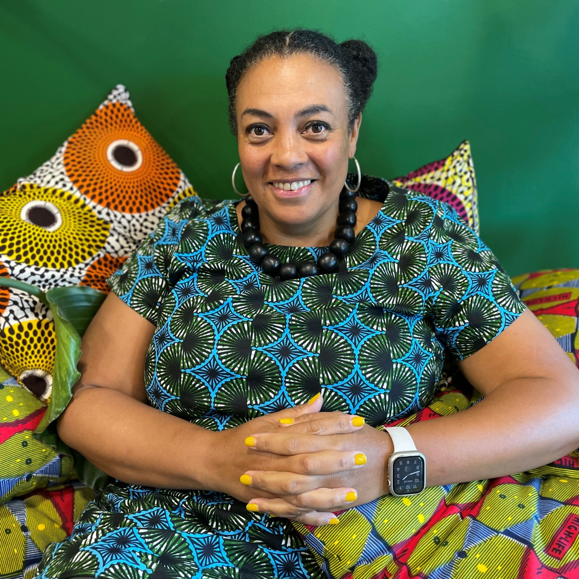 A woman with dark hair and a blue patterned dress, sitting down with arms crossed on a brightly coloured patterned sofa